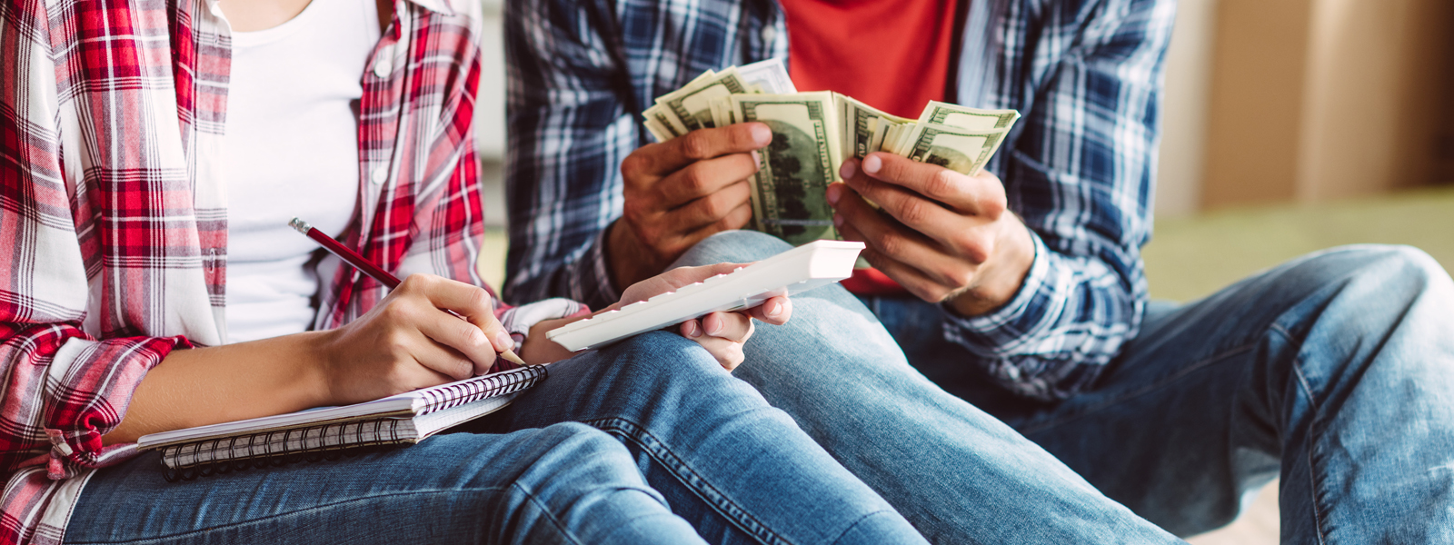 A man holding out cash while sitting next to a woman that is using a calculator and writing in a notebook.