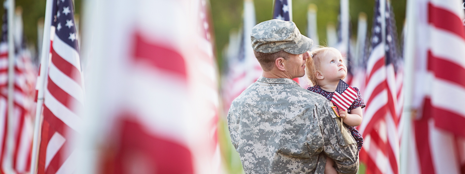 A Dad in an army uniform holding his daughter in a field of American flags.